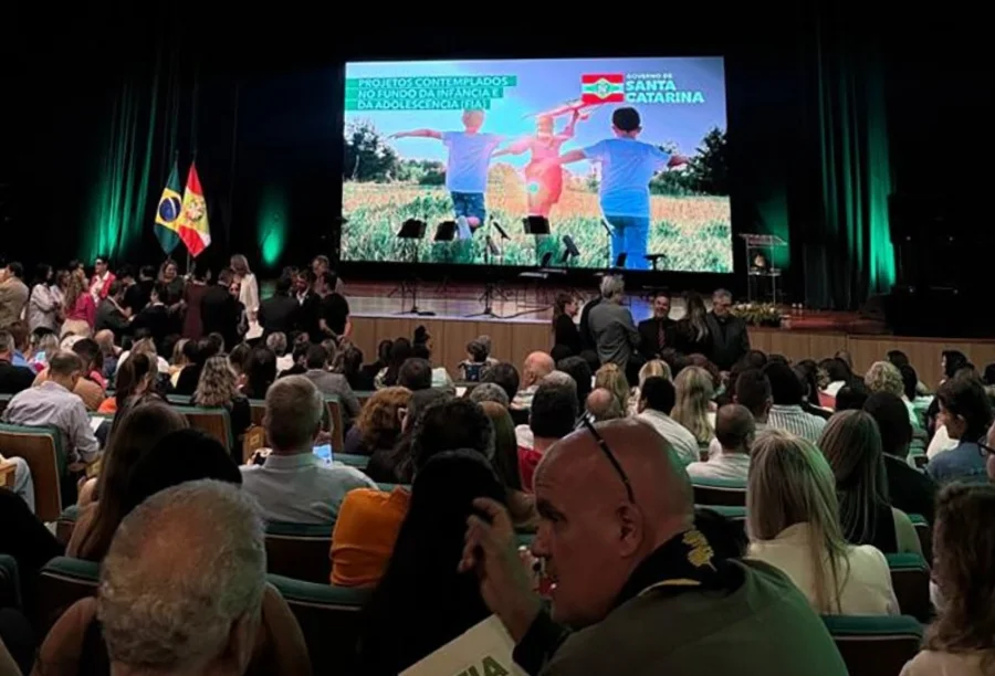 Santa Rosa do Sul garante R$ 676 mil para projeto de idosos Plenária lotada em evento com autoridades no palco, onde uma tela de LED exibe a mensagem