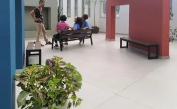 Saúde: Cuidados Paliativos serão levados à Atenção Primária Três pessoas conversando em uma área de convivência interna de uma unidade de saúde, com paredes nas cores azul e terracota. Um homem está em pé dialogando com duas mulheres vestidas de azul sentadas em um banco de madeira.