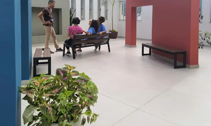 Saúde Cuidados Paliativos serão levados à Atenção Primária Três pessoas conversando em uma área de convivência interna de uma unidade de saúde, com paredes nas cores azul e terracota. Um homem está em pé dialogando com duas mulheres vestidas de azul sentadas em um banco de madeira.