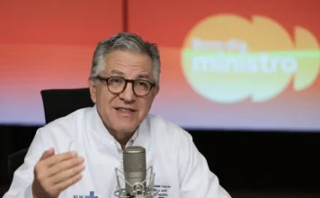 Saúde: Ministério processará quem lucra com fake news vacinal O Ministro da Saúde, Alexandre Padilha, de cabelos grisalhos e óculos de aro escuro, veste um jaleco branco e fala em um microfone de estúdio. Ele gesticula com a mão direita durante uma entrevista. Ao fundo, um painel em tons de laranja e amarelo exibe a frase 'Bom dia, Ministro'.