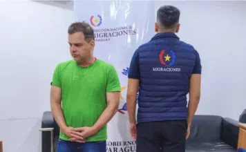 Silvinei Vasques tentou fugir para El Salvador com Pitbull Fotografia de Silvinei Vasques de pé, vestindo camiseta verde e calça jeans, com as mãos cruzadas à frente do corpo. À sua direita, um agente paraguaio de costas veste um colete azul com a inscrição "Migraciones". Ao fundo, um banner da Direção Nacional de Migrações do Paraguai. Ilustra sua detenção em solo estrangeiro.