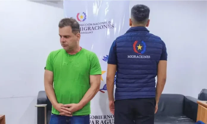 Silvinei Vasques tentou fugir para El Salvador com Pitbull Fotografia de Silvinei Vasques de pé, vestindo camiseta verde e calça jeans, com as mãos cruzadas à frente do corpo. À sua direita, um agente paraguaio de costas veste um colete azul com a inscrição "Migraciones". Ao fundo, um banner da Direção Nacional de Migrações do Paraguai. Ilustra sua detenção em solo estrangeiro.