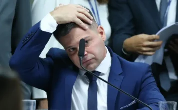 Silvinei tentou fugir do país com passaporte falso Close-up do ex-diretor da PRF Silvinei Vasques em um depoimento, vestindo terno azul e com a mão esquerda sobre a cabeça em sinal de preocupação.