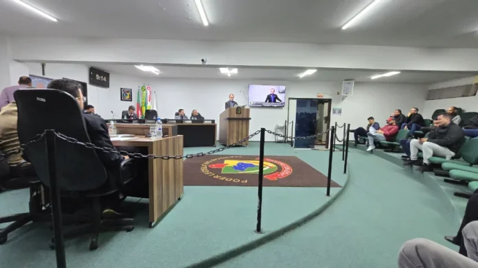 Sombrio Câmara aprova Censo da Pessoa com Deficiência Vista interna de um plenário da Câmara Municipal de Vereadores de Sombrio durante uma sessão. Um homem de terno está em pé, discursando em uma tribuna de madeira ao centro, com um microfone. Atrás dele, em uma tela de televisão, a imagem dele é repetida. No chão, um tapete circular escuro exibe o brasão da cidade e as palavras "PODER LEGISLATIVO MUNICIPAL". Vereadores estão sentados em mesas à esquerda, e o público assiste nas cadeiras verdes inclinadas à direita. A imagem registra a aprovação unânime do Censo Municipal das Pessoas com Deficiência.