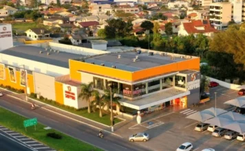 Sombrio Moda Shopping lança crediário com prazos estendidos Vista aérea do Sombrio Moda Shopping, um prédio branco e laranja com estacionamento, localizado próximo a uma avenida.