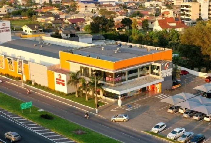 Sombrio Moda Shopping lança crediário com prazos estendidos Vista aérea do Sombrio Moda Shopping, um prédio branco e laranja com estacionamento, localizado próximo a uma avenida.