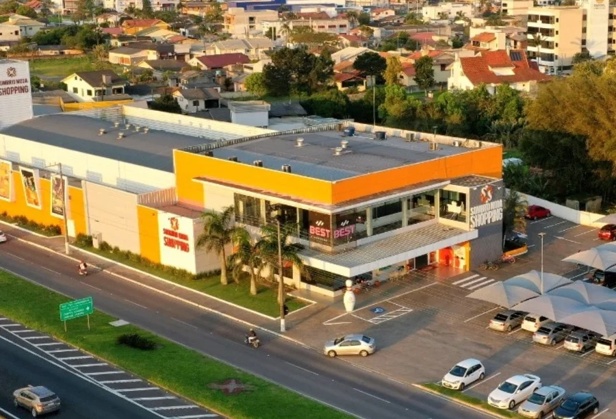 Sombrio Moda Shopping lança crediário com prazos estendidos Vista aérea do Sombrio Moda Shopping, um prédio branco e laranja com estacionamento, localizado próximo a uma avenida.