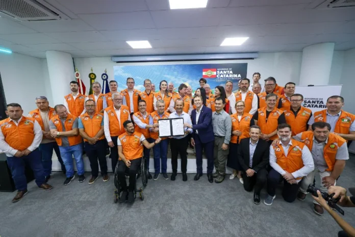 Sul de SC recebe R$ 102 milhões em investimento da Defesa Civil Governador Jorginho Mello e autoridades em coletes laranja da Defesa Civil posam segurando documentos, celebrando investimento de R$ 102 milhões.