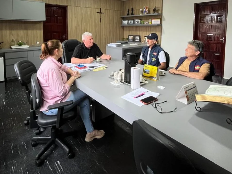 Timbé do Sul busca volta do transporte gratuito a Turvo Quatro pessoas reunidas em uma sala de escritório com móveis em tons de cinza e marrom escuro, sentadas ao redor de uma grande mesa cinza. A reunião inclui uma mulher (à esquerda, vestindo camisa rosa claro) e três homens (um de camisa polo preta e dois de boné e colete). Eles estão em um encontro para debater a volta do transporte gratuito para trabalhadores de Timbé do Sul até Turvo.