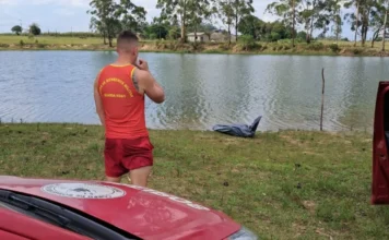 Tragédia em Sombrio: Jovem de 26 anos morre afogado em açude Membro do Corpo de Bombeiros Militar, vestindo camiseta vermelha de guarda-vidas e shorts vermelhos, em pé de costas para a câmera, olhando para a beira de um açude. Na grama, próximo à água, há um corpo coberto por um plástico preto. Em primeiro plano, é visível parte de uma viatura de resgate vermelha. A área é de mata, com árvores de eucalipto ao redor.