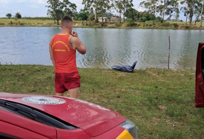 Tragédia em Sombrio Jovem de 26 anos morre afogado em açude Membro do Corpo de Bombeiros Militar, vestindo camiseta vermelha de guarda-vidas e shorts vermelhos, em pé de costas para a câmera, olhando para a beira de um açude. Na grama, próximo à água, há um corpo coberto por um plástico preto. Em primeiro plano, é visível parte de uma viatura de resgate vermelha. A área é de mata, com árvores de eucalipto ao redor.