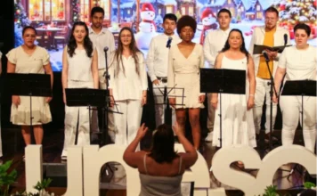 Unesc celebra fim de 2025 com foco na união e pertencimento Coral da Unesc composto por homens e mulheres vestidos de branco, apresentando-se em um palco. À frente, uma maestrina de costas rege o grupo. O cenário ao fundo exibe uma projeção natalina com bonecos de neve e vilas cobertas de neve. Letras grandes brancas formando o nome "Unesc" decoram a frente do palco.