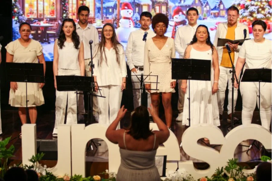 Coral da Unesc composto por homens e mulheres vestidos de branco, apresentando-se em um palco. À frente, uma maestrina de costas rege o grupo. O cenário ao fundo exibe uma projeção natalina com bonecos de neve e vilas cobertas de neve. Letras grandes brancas formando o nome 
