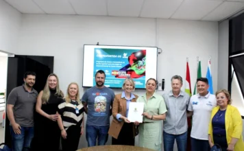 Unesc e Içara firmam parceria para contrapartida do UG Nove representantes da Unesc e da Prefeitura de Içara posam para foto em uma sala de reuniões. No centro, a reitora em exercício Gisele Lopes e a prefeita Dalvania Cardoso seguram juntas o documento oficial do termo de cooperação. Ao fundo, um monitor exibe um slide com o título "Contrapartida UG" e a bandeira de Santa Catarina.