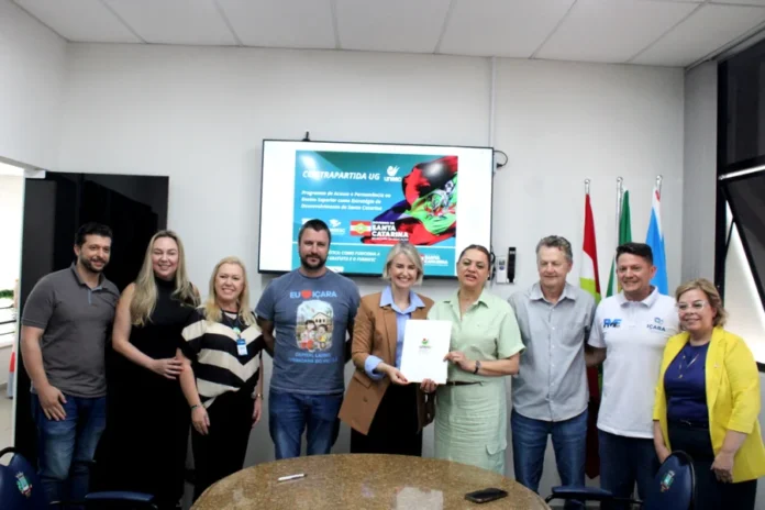 Unesc e Içara firmam parceria para contrapartida do UG Nove representantes da Unesc e da Prefeitura de Içara posam para foto em uma sala de reuniões. No centro, a reitora em exercício Gisele Lopes e a prefeita Dalvania Cardoso seguram juntas o documento oficial do termo de cooperação. Ao fundo, um monitor exibe um slide com o título "Contrapartida UG" e a bandeira de Santa Catarina.