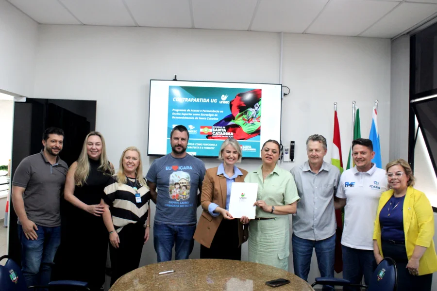 Unesc e Içara firmam parceria para contrapartida do UG Nove representantes da Unesc e da Prefeitura de Içara posam para foto em uma sala de reuniões. No centro, a reitora em exercício Gisele Lopes e a prefeita Dalvania Cardoso seguram juntas o documento oficial do termo de cooperação. Ao fundo, um monitor exibe um slide com o título