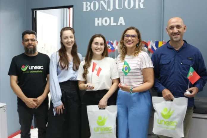 Unesc envia último grupo de intercâmbio de 2025 Grupo de cinco pesquisadores e acadêmicos da Unesc em pé, sorrindo e segurando pequenas bandeiras do Peru, Brasil e Portugal. Eles carregam sacolas brancas com o logotipo da universidade. Ao fundo, uma parede cinza com as palavras "BONJOUR" e "HOLA" escritas em destaque.