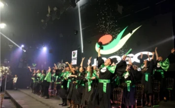 Unesc forma 40 odontólogos com foco em humanização e ciência Formandos de Odontologia da Unesc em becas pretas e faixas verdes celebram no palco com os braços erguidos e jogando seus capelos.