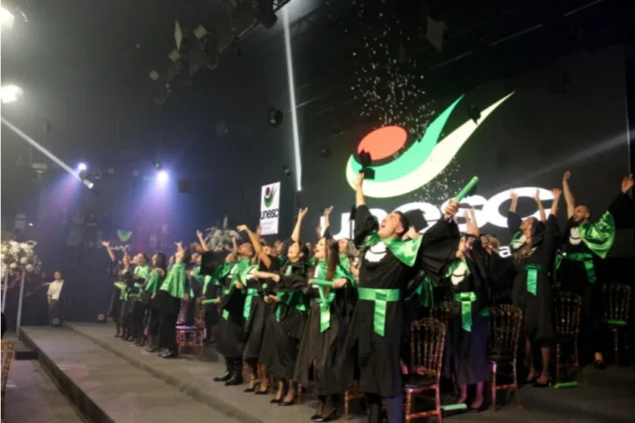 Unesc forma 40 odontólogos com foco em humanização e ciência Formandos de Odontologia da Unesc em becas pretas e faixas verdes celebram no palco com os braços erguidos e jogando seus capelos.