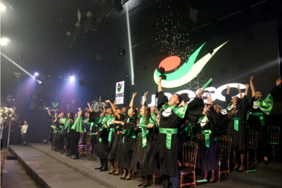 Unesc forma 40 odontólogos com foco em humanização e ciência Formandos de Odontologia da Unesc em becas pretas e faixas verdes celebram no palco com os braços erguidos e jogando seus capelos.