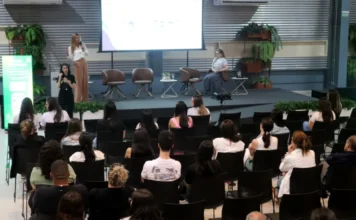 Unesc lança curso de Fonoaudiologia com palestra nacional Auditório do Centro de Inovação Criciúma (CRIO) durante o lançamento do curso de Fonoaudiologia da Unesc. No palco, a Dra. Ana Maria Furkim palestra em pé, enquanto uma intérprete de Libras realiza a tradução ao lado. O público, visto de costas, ocupa as cadeiras pretas do auditório, acompanhando a apresentação projetada em um grande telão ao fundo.