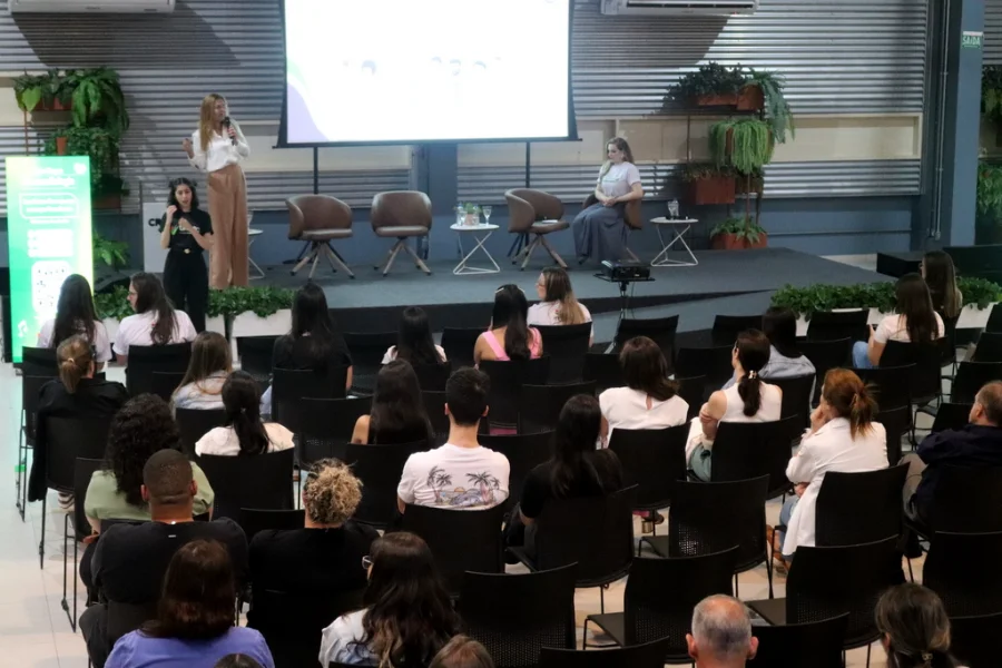 Auditório do Centro de Inovação Criciúma (CRIO) durante o lançamento do curso de Fonoaudiologia da Unesc. No palco, a Dra. Ana Maria Furkim palestra em pé, enquanto uma intérprete de Libras realiza a tradução ao lado. O público, visto de costas, ocupa as cadeiras pretas do auditório, acompanhando a apresentação projetada em um grande telão ao fundo.