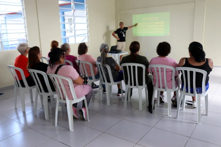Professor apresenta aula sobre exercício físico e alimentação saudável para um grupo de mulheres em um centro comunitário.