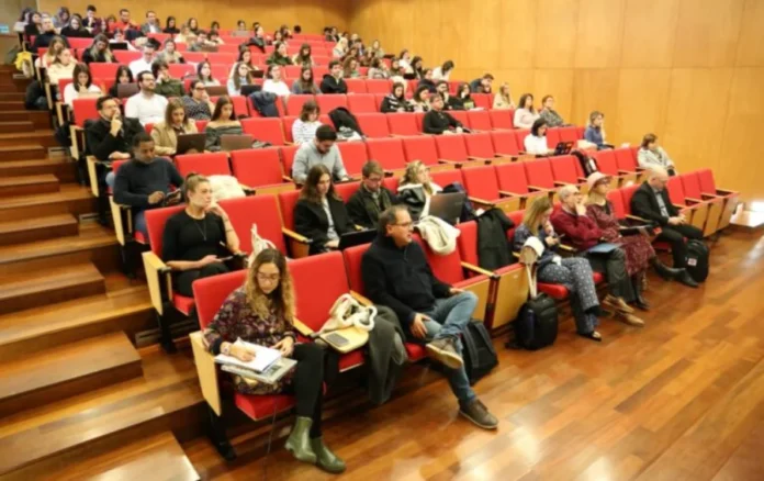Unesc representa Brasil em Portugal sobre IA e saúde mental Auditório em formato de anfiteatro na Universidade do Minho, em Portugal, lotado de participantes sentados em fileiras de cadeiras vermelhas em degraus de madeira. Vários participantes estão utilizando notebooks no colo e alguns fazem anotações, acompanhando a palestra sobre Direito e Tecnologias Digitais.