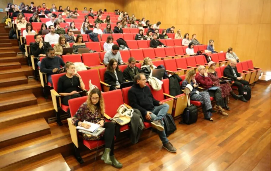 Unesc representa Brasil em Portugal sobre IA e saúde mental Auditório em formato de anfiteatro na Universidade do Minho, em Portugal, lotado de participantes sentados em fileiras de cadeiras vermelhas em degraus de madeira. Vários participantes estão utilizando notebooks no colo e alguns fazem anotações, acompanhando a palestra sobre Direito e Tecnologias Digitais.