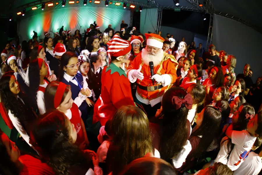 O Papai Noel, de microfone, abraça crianças e jovens coralistas no palco da Cantata de Luz da Unesc.