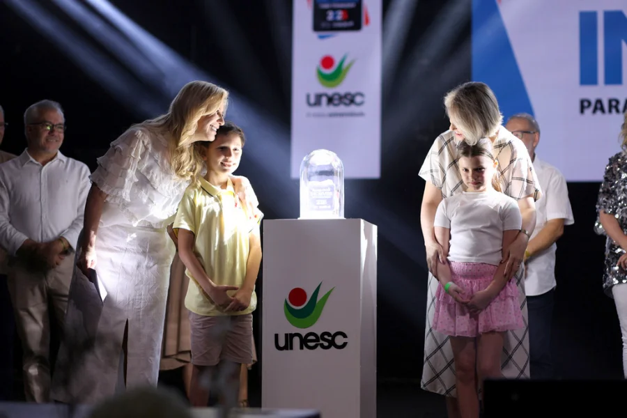 Reitoras e duas crianças em um palco iluminado, ao lado de um troféu de vidro sobre um pedestal com o logo da Unesc, após a premiação 