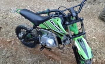 Urussanga: Adolescente cai de mini-moto ao fugir da PM Fotografia de uma mini-moto de 50 cilindradas, modelo TRX, nas cores verde e preta, caída sobre uma estrada de terra e brita. O veículo apresenta pneus de cravos sujos de lama e guidão preto. Ilustra a apreensão em Urussanga.