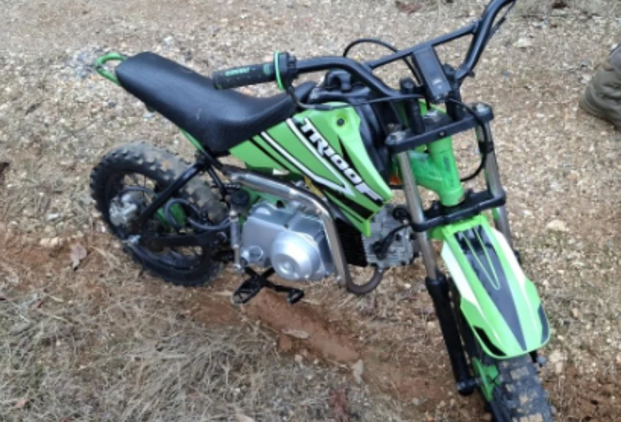 Fotografia de uma mini-moto de 50 cilindradas, modelo TRX, nas cores verde e preta, caída sobre uma estrada de terra e brita. O veículo apresenta pneus de cravos sujos de lama e guidão preto. Ilustra a apreensão em Urussanga.