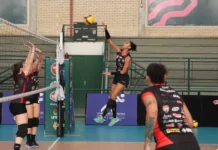 Vôlei de Criciúma faz estreia histórica na Superliga B Jogadora de vôlei do time Mampituba/Criciúma (camisa preta e vermelha) salta para cortar a bola por cima da rede, com duas adversárias bloqueando.