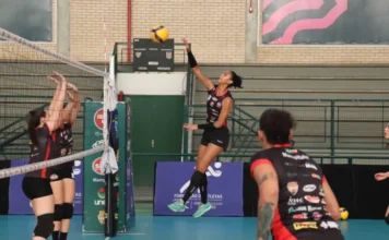 Vôlei de Criciúma faz estreia histórica na Superliga B Jogadora de vôlei do time Mampituba/Criciúma (camisa preta e vermelha) salta para cortar a bola por cima da rede, com duas adversárias bloqueando.