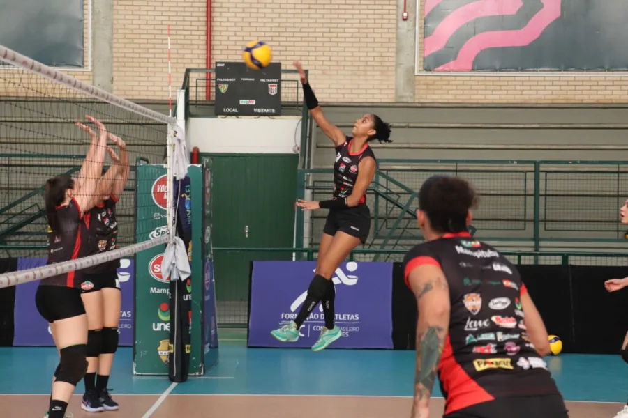 Jogadora de vôlei do time Mampituba/Criciúma (camisa preta e vermelha) salta para cortar a bola por cima da rede, com duas adversárias bloqueando.