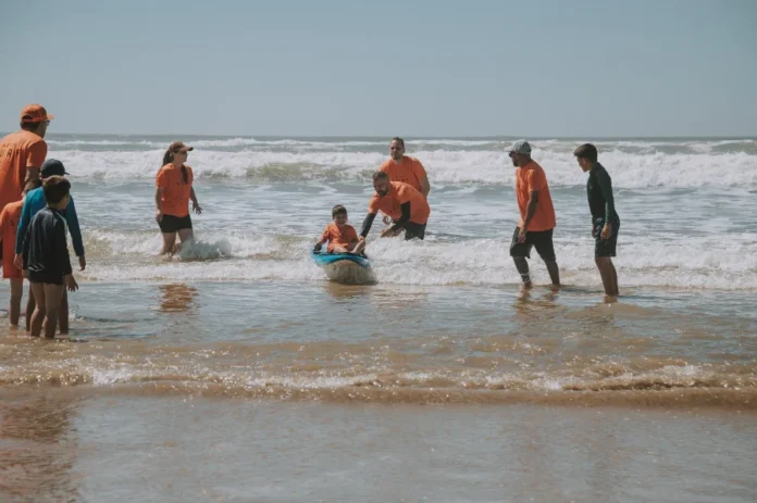 Abba Surf promove inclusão no Balneário Rincão neste sábado Grupo de voluntários do projeto Abba Surf, vestindo camisetas laranjas, auxiliam uma criança a surfar na beira do mar na praia do Balneário Rincão em um dia ensolarado.