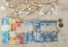 Adolescente é detido com crack no centro do Arroio do Silva Fotografia de cima para baixo exibindo o material apreendido: na parte superior, dezenas de pequenas pedras amareladas de crack individualmente embaladas em plástico transparente, organizadas em fileiras. Na parte inferior, notas de Real estendidas (uma de R$ 100, uma de R$ 20, uma de R$ 10 e quatro de R$ 2) ao lado de duas moedas, totalizando o valor de R$ 142,75 mencionado no relatório policial.