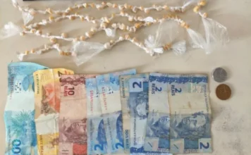 Adolescente é detido com crack no centro do Arroio do Silva Fotografia de cima para baixo exibindo o material apreendido: na parte superior, dezenas de pequenas pedras amareladas de crack individualmente embaladas em plástico transparente, organizadas em fileiras. Na parte inferior, notas de Real estendidas (uma de R$ 100, uma de R$ 20, uma de R$ 10 e quatro de R$ 2) ao lado de duas moedas, totalizando o valor de R$ 142,75 mencionado no relatório policial.