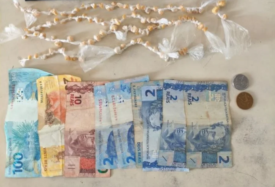 Fotografia de cima para baixo exibindo o material apreendido: na parte superior, dezenas de pequenas pedras amareladas de crack individualmente embaladas em plástico transparente, organizadas em fileiras. Na parte inferior, notas de Real estendidas (uma de R$ 100, uma de R$ 20, uma de R$ 10 e quatro de R$ 2) ao lado de duas moedas, totalizando o valor de R$ 142,75 mencionado no relatório policial.