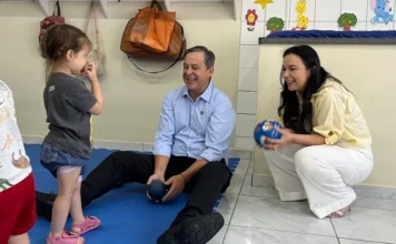 Afasc abre Colônia de Férias para crianças em Criciúma O prefeito de Criciúma, Vagner Espíndola, e a presidente de honra da Afasc, Gisele Bolan, brincam com uma criança sobre um tapete azul no CEI Lapagesse. O prefeito está sentado no chão e Gisele está agachada; ambos sorriem e seguram bolas coloridas, interagindo com a menina. Ao fundo, a parede exibe decorações infantis e mochilas penduradas.