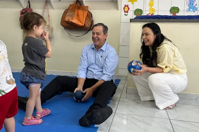 Afasc abre Colônia de Férias para crianças em Criciúma O prefeito de Criciúma, Vagner Espíndola, e a presidente de honra da Afasc, Gisele Bolan, brincam com uma criança sobre um tapete azul no CEI Lapagesse. O prefeito está sentado no chão e Gisele está agachada; ambos sorriem e seguram bolas coloridas, interagindo com a menina. Ao fundo, a parede exibe decorações infantis e mochilas penduradas.