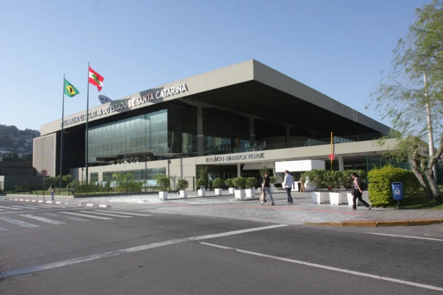 Fachada ensolarada do Palácio Barriga Verde, sede da Assembleia Legislativa de Santa Catarina (Alesc) em Florianópolis. O prédio moderno de vidro e concreto possui bandeiras hasteadas e o nome da instituição na frente, sendo o local de trabalho dos servidores que completam um ano de posse.