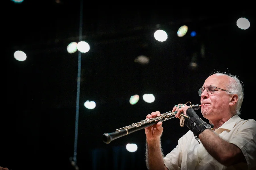 Maestro Alex Klein tocando oboé no palco com iluminação ao fundo; o músico será homenageado com o Título de Cidadão Catarinense.