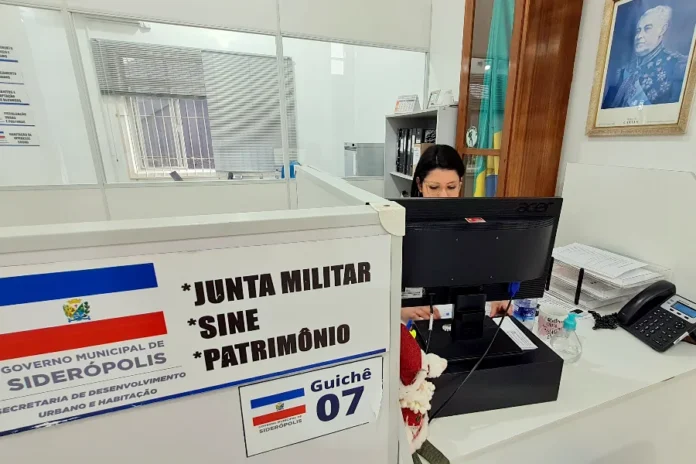 Alistamento Militar 2026 Siderópolis abre inscrições Uma funcionária trabalha em um computador atrás de um balcão de atendimento com a identificação "GUICHÊ 07" e "JUNTA MILITAR / SINE / PATRIMÔNIO" na Prefeitura de Siderópolis. Na parede, há um retrato emoldurado do Duque de Caxias.