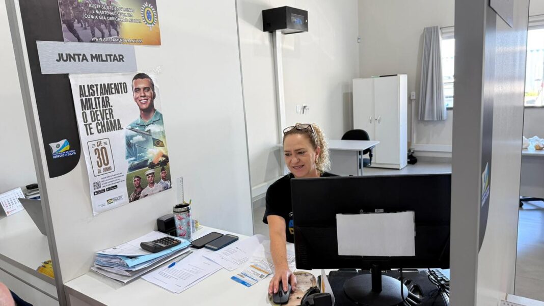 Atendimento na Junta de Serviço Militar de Balneário Arroio do Silva, com cartazes ao fundo informando o prazo final de 30 de junho para o alistamento militar.