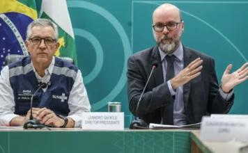 Anvisa libera testes com remédio nacional para lesão medular Fotografia de dois homens sentados à mesa em uma coletiva de imprensa. À esquerda, o ministro Alexandre Padilha, de colete azul do SUS sobre camisa listrada. À direita, o diretor da Anvisa, Leandro Safatle, de terno cinza e óculos, gesticula com as mãos enquanto fala. Ao fundo, uma parede verde com o brasão da República e a bandeira nacional.