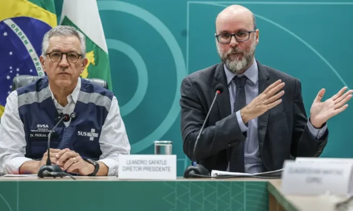 Anvisa libera testes com remédio nacional para lesão medular Fotografia de dois homens sentados à mesa em uma coletiva de imprensa. À esquerda, o ministro Alexandre Padilha, de colete azul do SUS sobre camisa listrada. À direita, o diretor da Anvisa, Leandro Safatle, de terno cinza e óculos, gesticula com as mãos enquanto fala. Ao fundo, uma parede verde com o brasão da República e a bandeira nacional.