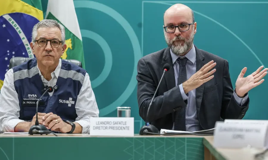 Anvisa libera testes com remédio nacional para lesão medular Fotografia de dois homens sentados à mesa em uma coletiva de imprensa. À esquerda, o ministro Alexandre Padilha, de colete azul do SUS sobre camisa listrada. À direita, o diretor da Anvisa, Leandro Safatle, de terno cinza e óculos, gesticula com as mãos enquanto fala. Ao fundo, uma parede verde com o brasão da República e a bandeira nacional.