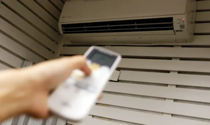 Ar-condicionado como usar sem pesar no bolso Mão segurando um controle remoto apontado para um ar-condicionado split na parede, ilustrando o ajuste da temperatura ideal para economizar energia.